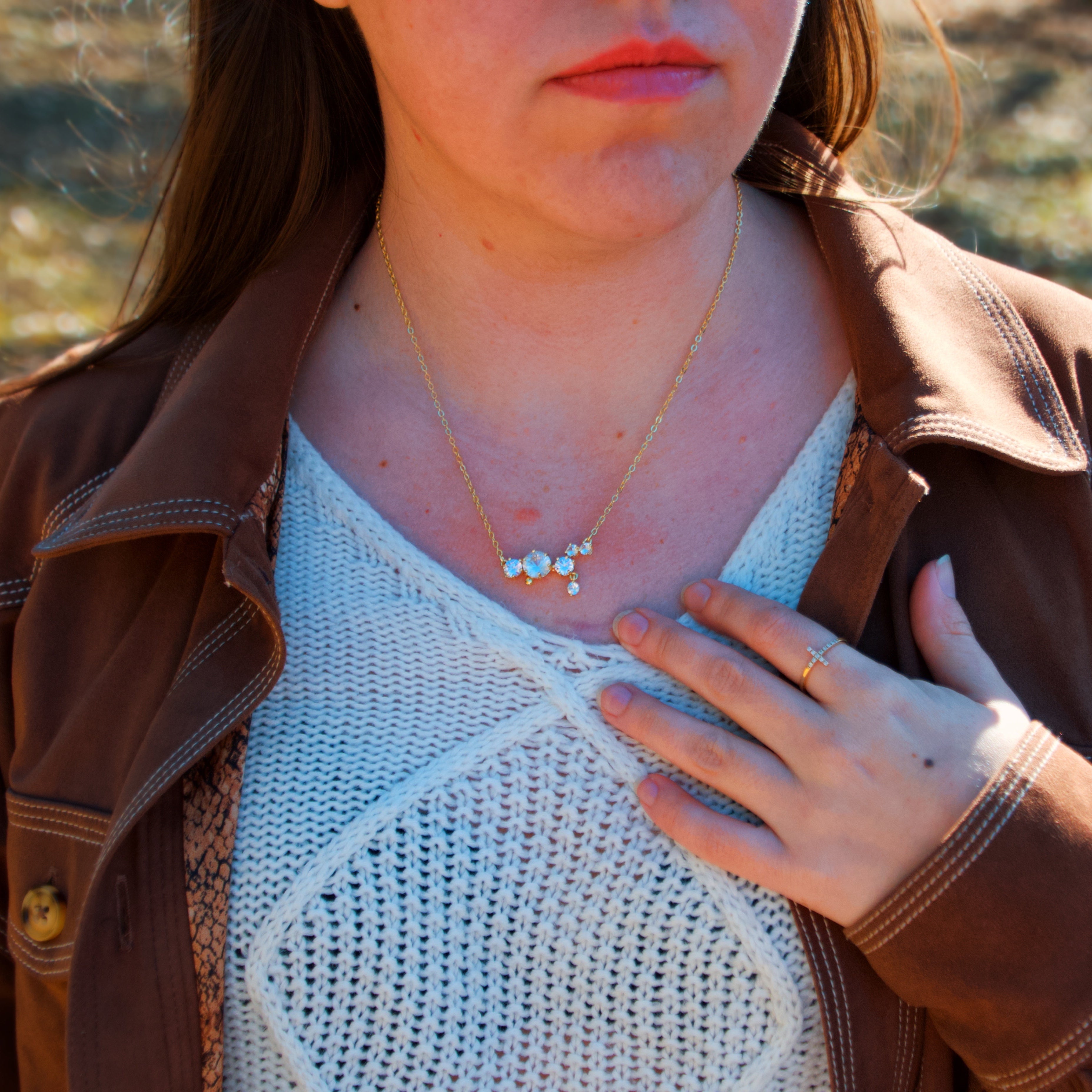 A close-up lifestyle shot of a woman wearing a delicate gold necklace with a small cluster of white stones, styled with a brown jacket and white knit top outdoors.