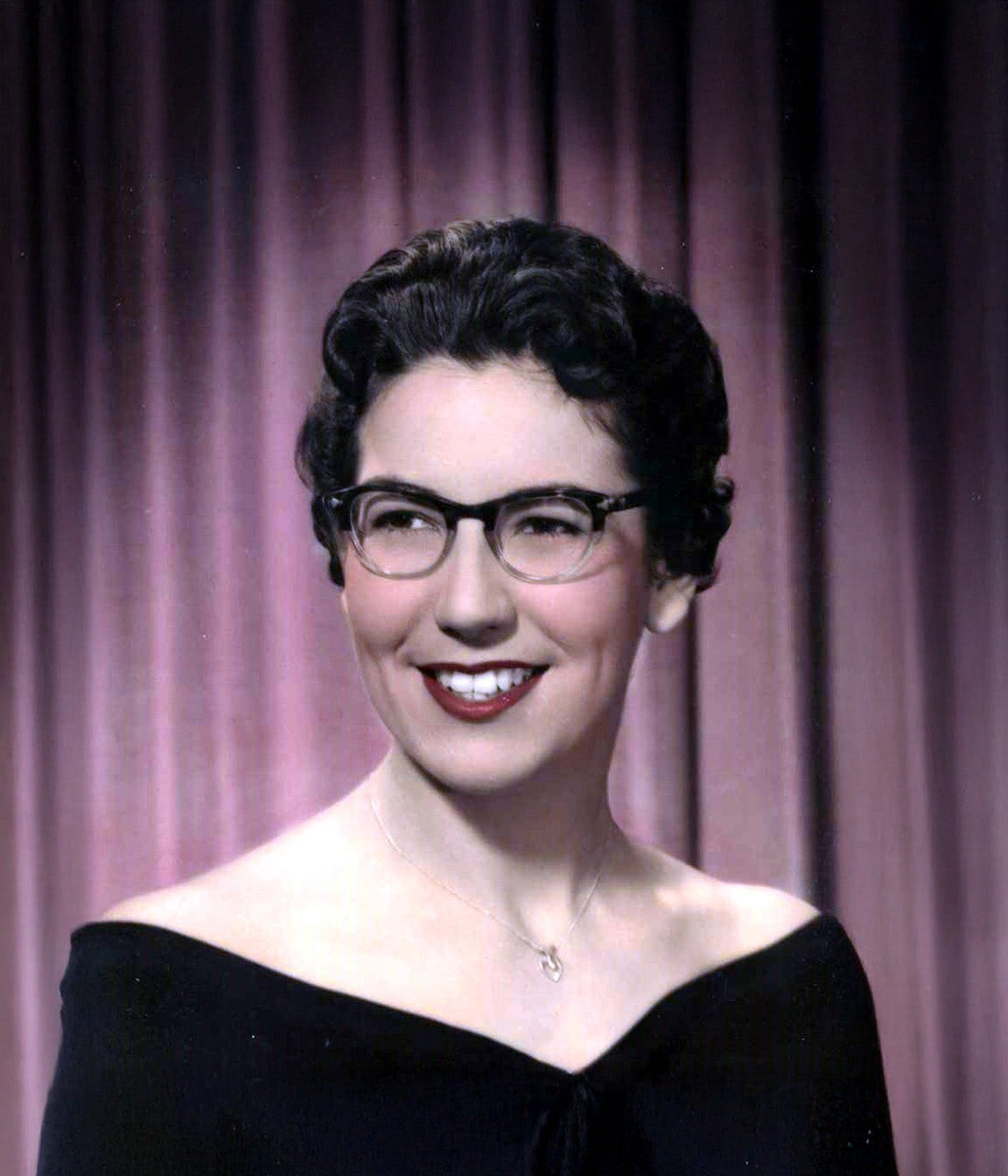 Woman wearing glasses and a black top against a purple curtain background