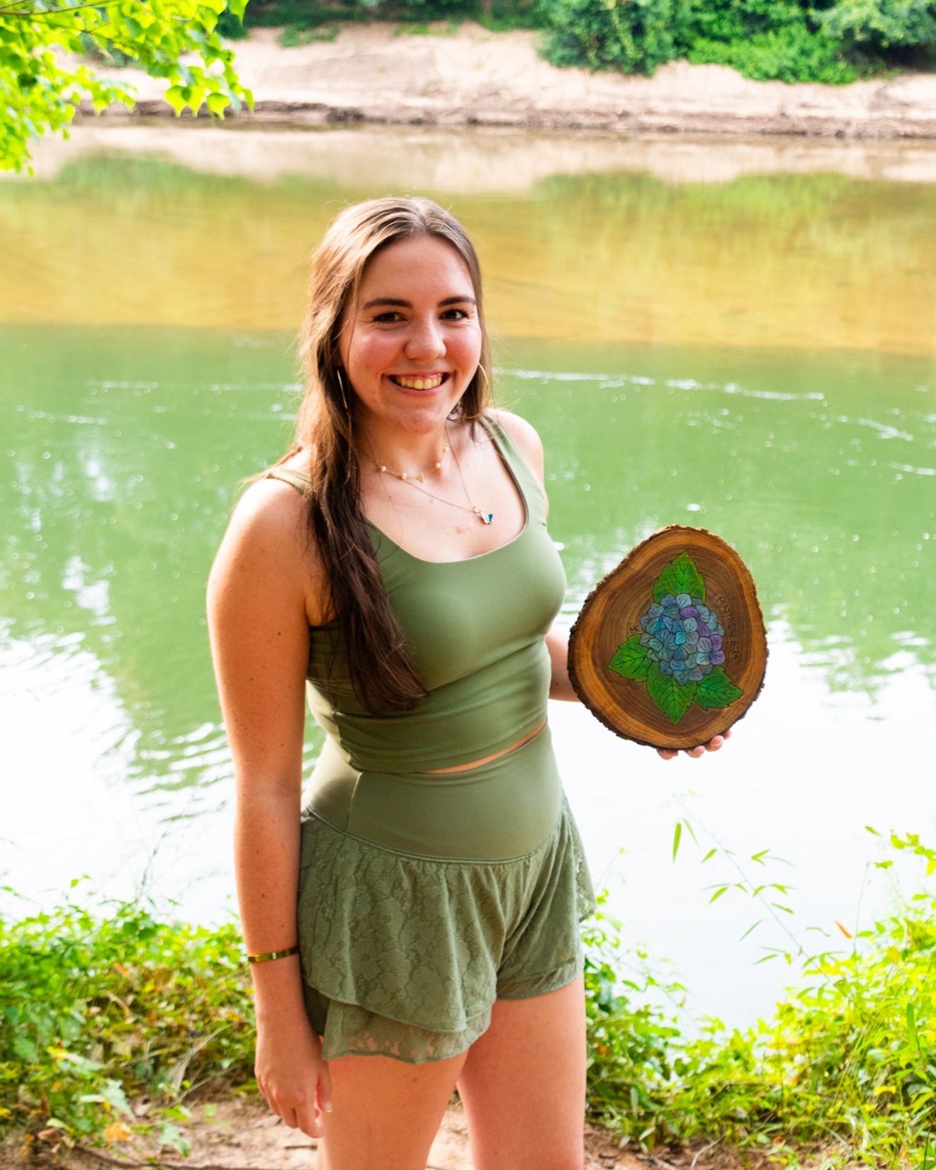 Woman holding a wooden disc with a floral design by a body of water
